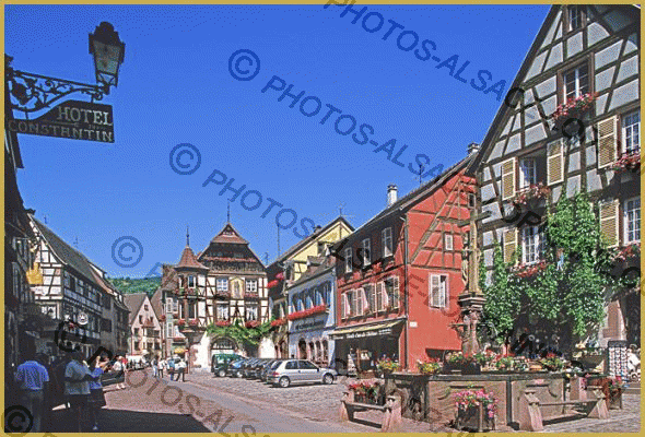 Photo de la rue du Général de Gaulle bordée de belles maisons alsaciennes à pans de bois en fleurs, avec la fontaine Constantin, dans le centre historique de Kaysersberg au mois de juillet. Description et visite de Kaysersberg.