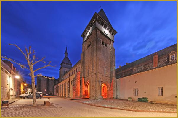 Photo de l'abbatiale Sainte-Valérie illuminée à la tombée de la nuit, un des plus importants édifices de style roman du Limousin, avec son clocher-porche dit ''tour des bourgeois'', et sa grande tour-clocher dite ''tour du Chartrier''.